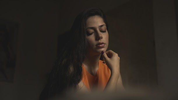 A woman with long dark hair is sitting with her eyes closed, appearing deep in thought or contemplation. She wears an orange top, and her hand is gently touching her chin. The lighting is dim, casting a soft, moody shadow around her.