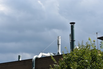 Several industrial chimneys and vents rise from the roof of a building against a backdrop of overcast gray clouds. Some green foliage is visible in the foreground.