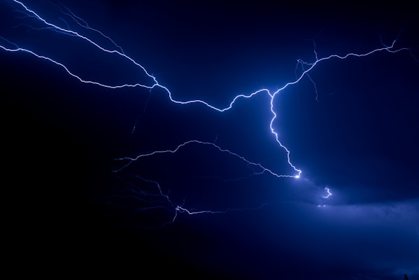 Intense lightning bolts illuminate a dark night sky, with bright flashes cutting through thick, ominous clouds. The scene is dominated by dark blue and deep black tones, highlighting the raw power of the natural phenomenon.