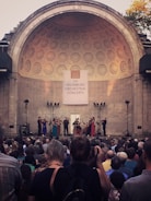 A large audience is gathered in front of an orchestral performance, set against the backdrop of an architecturally detailed amphitheater. Musicians, dressed in formal attire, are positioned under soft stage lighting, playing string instruments. The atmosphere is serene, with focus lights directed at the musicians.