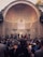 A large audience is gathered in front of an orchestral performance, set against the backdrop of an architecturally detailed amphitheater. Musicians, dressed in formal attire, are positioned under soft stage lighting, playing string instruments. The atmosphere is serene, with focus lights directed at the musicians.