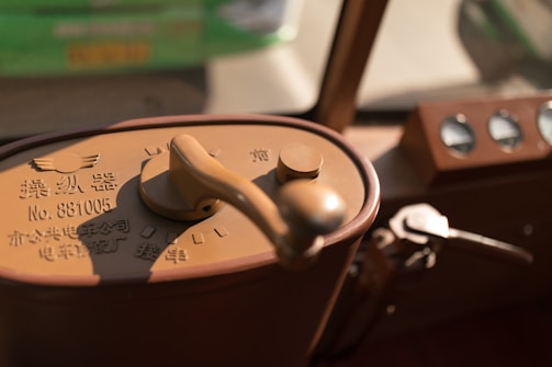 A close-up view of a vintage machine control panel with a large handle and various dials. The control panel has Chinese characters and a serial number imprinted on it, with a curved metallic lever visible beside it. The background is out of focus, revealing a mix of green and beige colors.