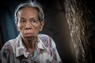 An elderly person with a contemplative expression is captured in a seated position, wearing a patterned shirt with a mix of pastel colors. The background is out of focus, featuring textured surfaces and dim lighting.