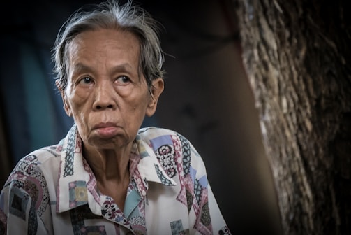 An elderly person with a contemplative expression is captured in a seated position, wearing a patterned shirt with a mix of pastel colors. The background is out of focus, featuring textured surfaces and dim lighting.
