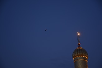 A mosque dome with a crescent moon symbol is lit up against a dark blue evening sky. A drone is seen flying above, its silhouette barely visible.