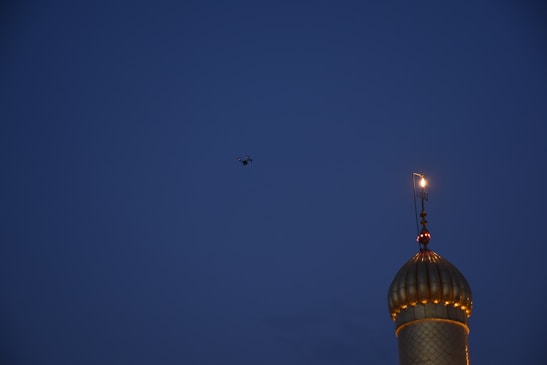 A mosque dome with a crescent moon symbol is lit up against a dark blue evening sky. A drone is seen flying above, its silhouette barely visible.