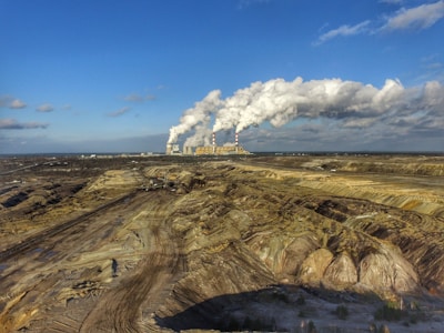 An industrial landscape featuring a large power plant in the distance emitting thick plumes of smoke from multiple chimneys. The foreground consists of a barren, excavated land, possibly a mining site, with visible tracks and machinery lines. The sky is mostly clear with some scattered clouds.