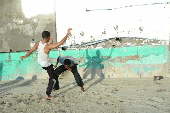 Two individuals are engaged in a form of martial arts or dance on a sandy surface against a backdrop of a weathered and peeling wall. One person is in a white tank top and black pants, performing a dynamic movement, while the other is in a bent posture, appearing to brace against the action. Shadows are cast on the wall, adding a sense of motion.