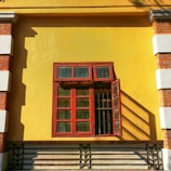A closed decorative red wooden window with glass panels is mounted on a vibrant yellow wall. The window contains four larger panes below and three smaller panes above. One of the smaller panes on the right side is open, casting long shadows. The wall features bricks and white plaster decorations on the corners, adding a rustic and vintage touch.