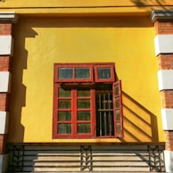 A closed decorative red wooden window with glass panels is mounted on a vibrant yellow wall. The window contains four larger panes below and three smaller panes above. One of the smaller panes on the right side is open, casting long shadows. The wall features bricks and white plaster decorations on the corners, adding a rustic and vintage touch.