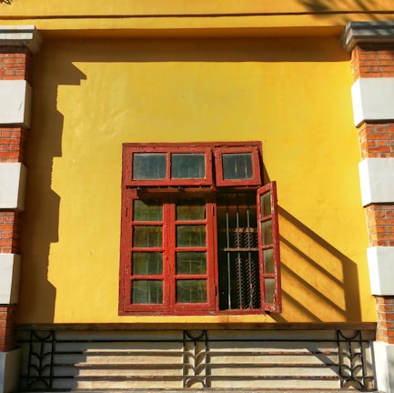 A closed decorative red wooden window with glass panels is mounted on a vibrant yellow wall. The window contains four larger panes below and three smaller panes above. One of the smaller panes on the right side is open, casting long shadows. The wall features bricks and white plaster decorations on the corners, adding a rustic and vintage touch.