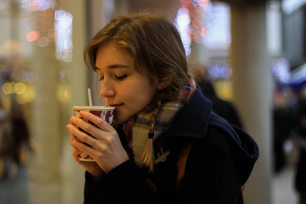 A person enjoying a warm drink from a paper cup, closed eyes suggest a moment of contentment or relaxation. They are wearing a scarf and coat, indicating a cold environment. The background contains blurred lights, possibly indicating a festive or urban setting.