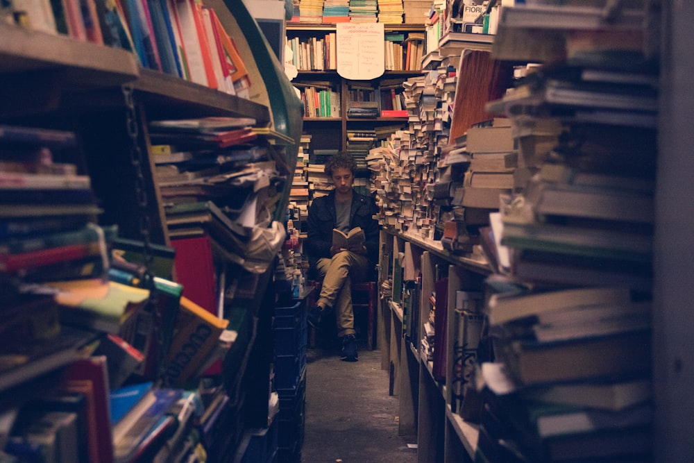 Mann sitzt Buch lesend auf dem Ende eines Stapels von Bücherregalen