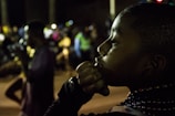 A young boy is seen in profile, his hand resting near his mouth as if in deep thought. The background is blurred with several people, hinting at a busy or social setting, possibly during nighttime. Some individuals in the background appear to be engaged in a performance or procession.