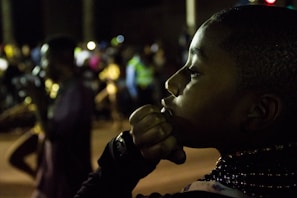 A young boy is seen in profile, his hand resting near his mouth as if in deep thought. The background is blurred with several people, hinting at a busy or social setting, possibly during nighttime. Some individuals in the background appear to be engaged in a performance or procession.