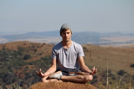 A person is meditating in a seated position with crossed legs on a rock, surrounded by a natural landscape with hills and a clear sky in the background. The individual is wearing a gray t-shirt, shorts, a beanie, and has a relaxed posture with hands resting on their knees.