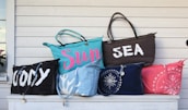 A collection of six colorful beach bags displayed on a white wooden surface. The bags have various inscriptions and designs. One bag has the word 'WODY' in large white letters, another reads 'Sun' in pink on a blue bag, and another features 'SEA' in white on a brown bag. Other bags have patterns resembling coral and seashells.