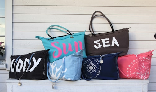 A collection of six colorful beach bags displayed on a white wooden surface. The bags have various inscriptions and designs. One bag has the word 'WODY' in large white letters, another reads 'Sun' in pink on a blue bag, and another features 'SEA' in white on a brown bag. Other bags have patterns resembling coral and seashells.