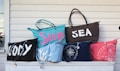 A collection of six colorful beach bags displayed on a white wooden surface. The bags have various inscriptions and designs. One bag has the word 'WODY' in large white letters, another reads 'Sun' in pink on a blue bag, and another features 'SEA' in white on a brown bag. Other bags have patterns resembling coral and seashells.