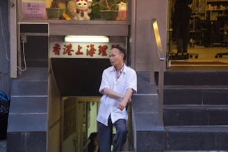 A man in a white shirt stands casually at the entrance of a shop, leaning against the stair railing. Above him, a sign with Chinese characters is visible, and inside the shop, a figure can be seen along with shelves and various items, including a waving cat figurine.