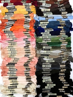 Multiple rows of colorful embroidery threads are neatly arranged. The threads are organized by color, ranging across different shades including red, orange, yellow, pink, green, blue, and brown. Each thread has a label with brand and color information.