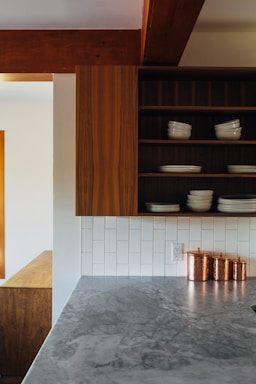 A modern kitchen with a gray marble countertop, featuring a wooden cabinet with open shelves displaying white dishes. The backsplash consists of white rectangular tiles, and there are three copper storage canisters on the counter.