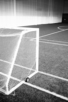 A monochrome photograph of an indoor sports field featuring a small wheeled goalpost on artificial turf. The goal's white netting stands out against the dark background of the field. The image captures the lines of the sports field, which add to the geometric composition.