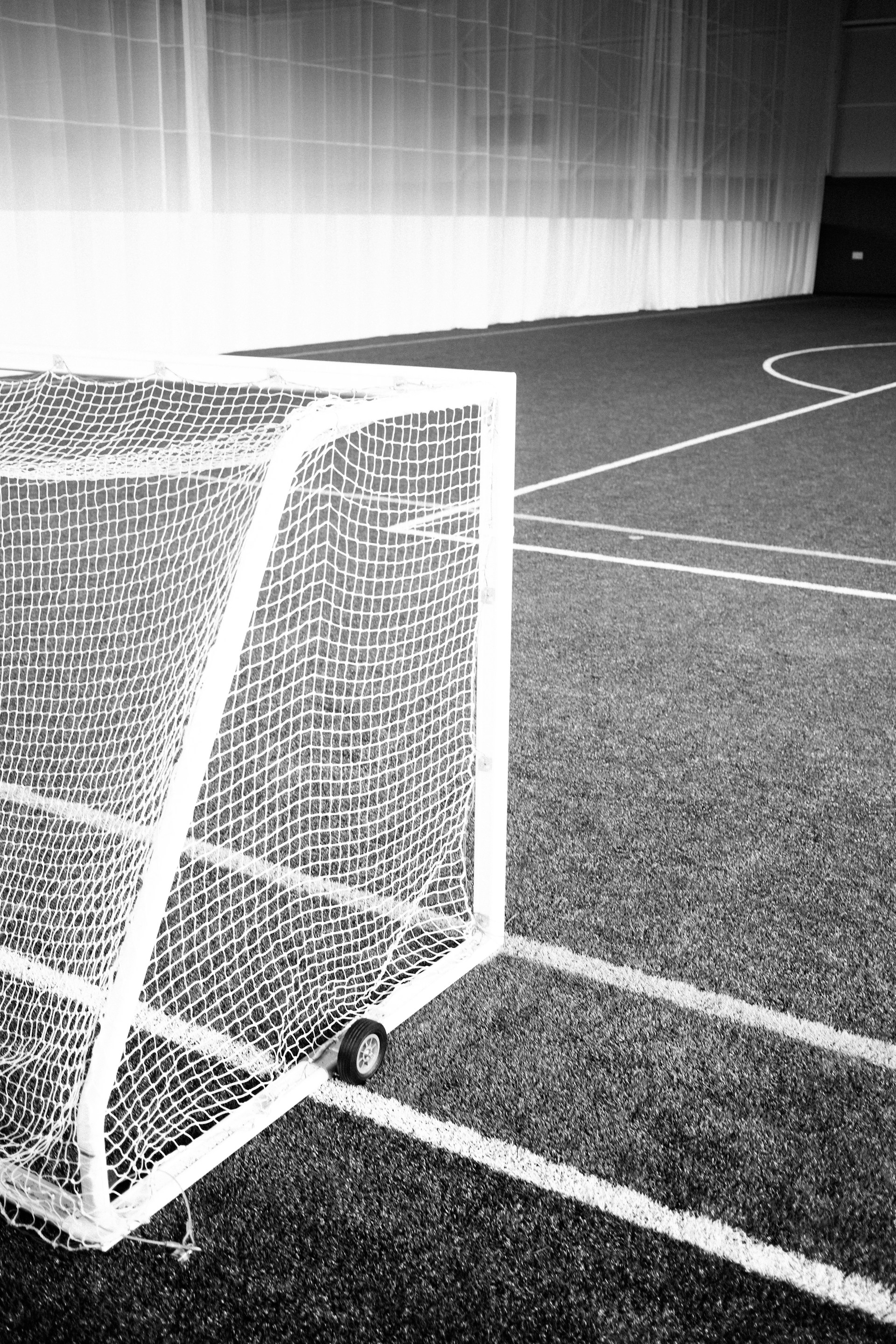 A monochrome photograph of an indoor sports field featuring a small wheeled goalpost on artificial turf. The goal's white netting stands out against the dark background of the field. The image captures the lines of the sports field, which add to the geometric composition.