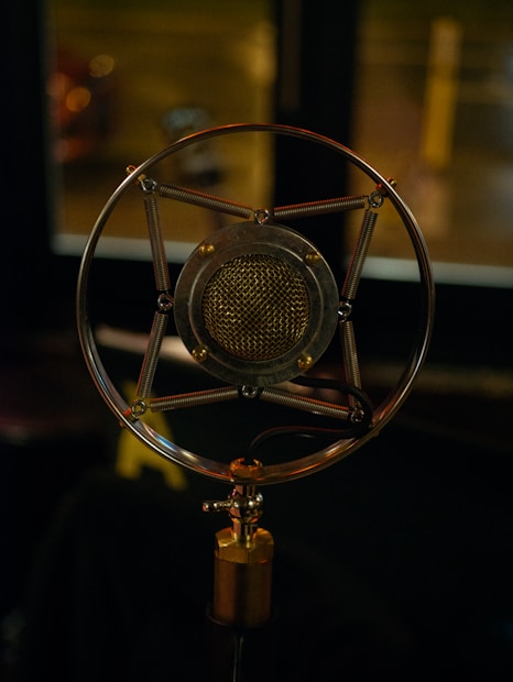 An antique-style microphone with a round metallic frame and a mesh grill is positioned against a softly lit background. The intricate design features metal springs and a central mesh component, evoking a vintage appearance.