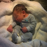 A newborn baby is peacefully sleeping, dressed in a grey outfit with small patterns. The baby is resting on a soft, white blanket, creating a cozy and serene environment.
