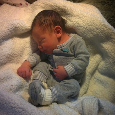 A newborn baby is peacefully sleeping, dressed in a grey outfit with small patterns. The baby is resting on a soft, white blanket, creating a cozy and serene environment.