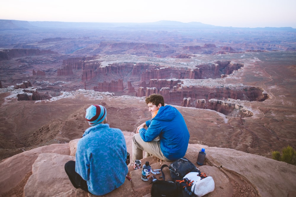 two man sitting on the edge of a cliff during daytime