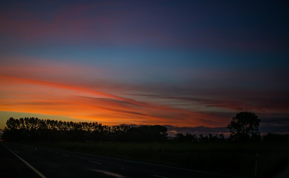 The image displays a tranquil sunset scene with vibrant hues of orange and pink in the sky, transitioning to deep blue as the night approaches. Silhouettes of trees line the horizon, creating a serene and peaceful landscape.