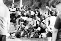 A group of rugby players engage in a scrum during a match on a grassy field. Intensity and focus are evident as they compete for possession of the ball. Fans and fellow players surround the scene, adding to the dynamic atmosphere.