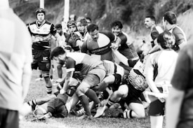 A group of rugby players engage in a scrum during a match on a grassy field. Intensity and focus are evident as they compete for possession of the ball. Fans and fellow players surround the scene, adding to the dynamic atmosphere.