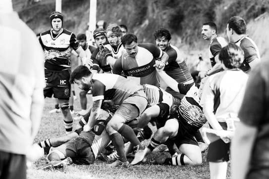 A group of rugby players engage in a scrum during a match on a grassy field. Intensity and focus are evident as they compete for possession of the ball. Fans and fellow players surround the scene, adding to the dynamic atmosphere.