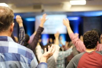 A group of people with raised hands in what appears to be a conference or meeting room. The focus is on the backs of individuals, as they face a stage or screen with blurred content, creating an atmosphere of engagement or participation.