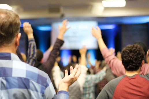 A group of people with raised hands in what appears to be a conference or meeting room. The focus is on the backs of individuals, as they face a stage or screen with blurred content, creating an atmosphere of engagement or participation.