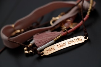 A close-up of a bracelet with a gold-colored rectangular plate engraved with the words ‘Make Today Amazing’. The bracelet is adorned with various beads and a tassel in soft pink and brown tones, laid out on a dark surface.