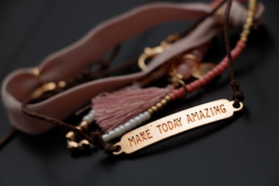 A close-up of a bracelet with a gold-colored rectangular plate engraved with the words ‘Make Today Amazing’. The bracelet is adorned with various beads and a tassel in soft pink and brown tones, laid out on a dark surface.