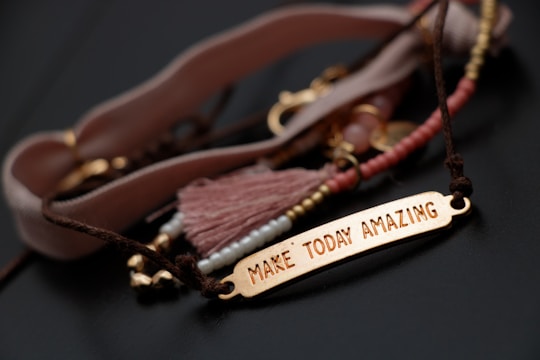 A close-up of a bracelet with a gold-colored rectangular plate engraved with the words ‘Make Today Amazing’. The bracelet is adorned with various beads and a tassel in soft pink and brown tones, laid out on a dark surface.