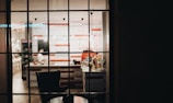 A view through a window into a cozy cafe or restaurant with a counter and stools. The interior is warmly lit with hanging pendant lights and features a striped wall design in red and white. Various items like bottles and plants are visible on the counter.