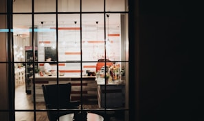 A view through a window into a cozy cafe or restaurant with a counter and stools. The interior is warmly lit with hanging pendant lights and features a striped wall design in red and white. Various items like bottles and plants are visible on the counter.