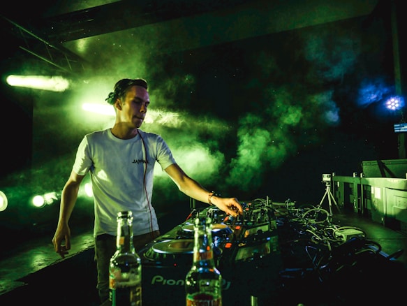 A DJ wearing a white t-shirt is performing on stage, surrounded by smoke and green lighting effects. The setup includes audio equipment with cables and a camera on a tripod in the background. Bottles are visible on a table in the foreground.