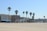 A sandy beach with multiple palm trees lining the edge. Behind the trees, there are several multi-story buildings with a mix of architectural styles. Some structures have blue accents and awnings, and there are people visible along the walkway in front of the buildings. The sky is clear and light blue.