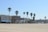 A sandy beach with multiple palm trees lining the edge. Behind the trees, there are several multi-story buildings with a mix of architectural styles. Some structures have blue accents and awnings, and there are people visible along the walkway in front of the buildings. The sky is clear and light blue.