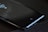 A smartphone lies on a table with its screen displaying a small square icon near the bottom. The surface is mostly black with subtle reflections and hints of light.