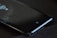 A smartphone lies on a table with its screen displaying a small square icon near the bottom. The surface is mostly black with subtle reflections and hints of light.