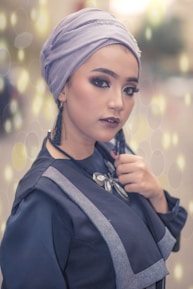A person is wearing a light purple headscarf and dark makeup, including eyeliner and lipstick. The person is also wearing dark clothing with gray accents and a decorative necklace. The background features a soft bokeh effect with circular light patterns.