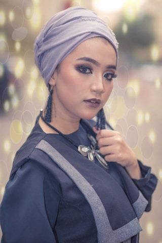 A person is wearing a light purple headscarf and dark makeup, including eyeliner and lipstick. The person is also wearing dark clothing with gray accents and a decorative necklace. The background features a soft bokeh effect with circular light patterns.